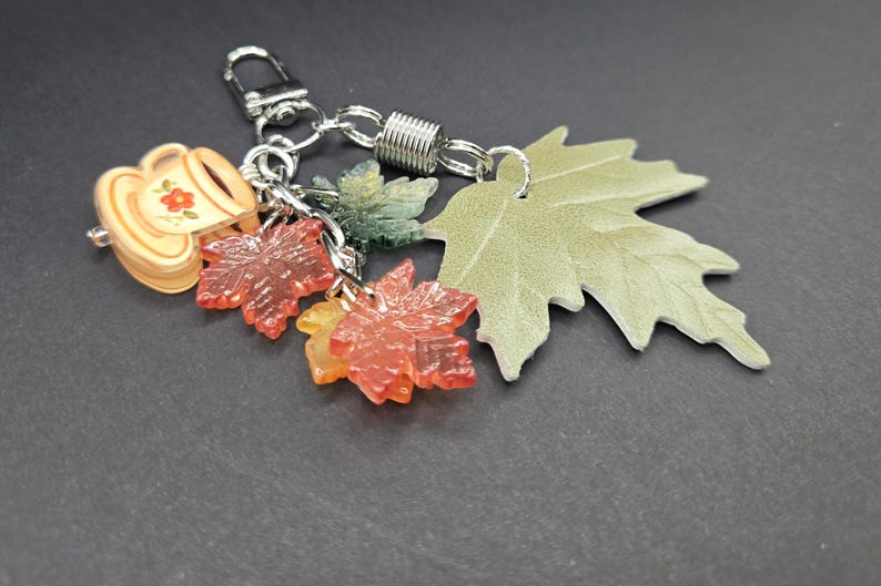 May include: A silver-toned metal keychain featuring a collection of charms including a small beige cup with a red flower design, two translucent red maple leaf charms, one dark green leaf charm, and a larger light green textured maple leaf.