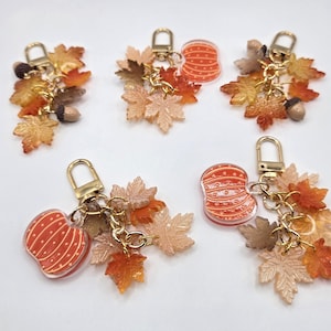 May include: Five keychains featuring gold-tone lobster clasps, each adorned with various resin charms including orange pumpkins with white stripes, brown acorns, and multiple maple leaves in shades of yellow, orange, and brown, arranged in clusters.