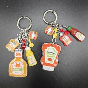 May include: Two keychains with silver rings, each adorned with food-themed charms. One features a ketchup bottle, a hotdog, and a fork. The other has a mustard bottle, a sauce bottle, and a small spoon. The charms are colorful and cartoonish.