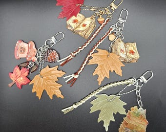 May include: Three keychains featuring autumn-themed charms including maple leaves in red, tan, and pale green, miniature open books with leaf illustrations, small acorns, a pinecone, and a small house charm, all attached to silver-toned metal clasps.