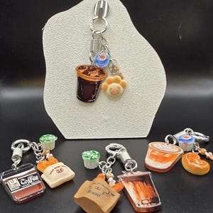 May include: A collection of keychains featuring miniature food and drink charms. The keychains include charms of iced coffee, bread, and pastries. The keychains are attached to silver-colored metal rings and clasps. Some charms have a cartoon-like design.