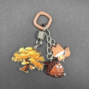 May include: A keychain featuring a light brown square carabiner attached to a silver metal spring ring. Three charms hang from the ring: a flat acrylic tree with yellow and orange autumn leaves, a small orange fox, and a brown textured acorn.