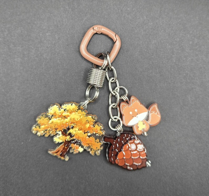 May include: A keychain featuring a light brown square carabiner attached to a silver metal spring ring. Three charms hang from the ring: a flat acrylic tree with yellow and orange autumn leaves, a small orange fox, and a brown textured acorn.