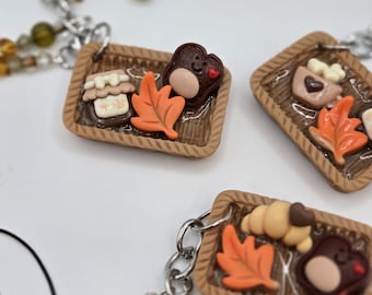 Könnte beinhalten: Nahaufnahme von drei Miniatur-Tablett-Anhängern mit herbstlichen Motiven. Jedes Tablett hat ein braunes Korbgeflecht-Design und enthält ein Ahornblatt, einen Bären und andere Lebensmittel. Die Anhänger sind an einer silbernen Kette mit dekorativen Perlen befestigt.