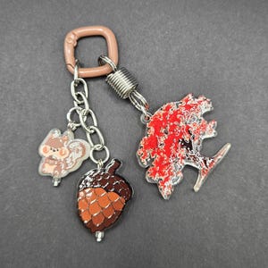 May include: A keychain featuring a light brown square clasp, a silver chain, and three charms: a small light brown squirrel, a brown acorn with a textured cap, and a larger charm shaped like a tree with vibrant red and white foliage.