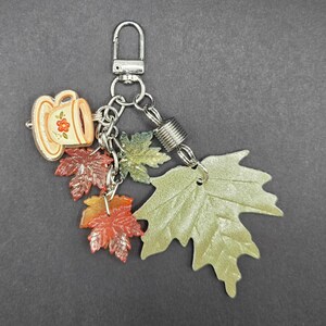 May include: A silver-toned metal keychain featuring a small cream-colored teacup charm with a red floral design, accompanied by several dangling maple leaf charms in shades of light green, dark red, and orange, attached by silver jump rings.