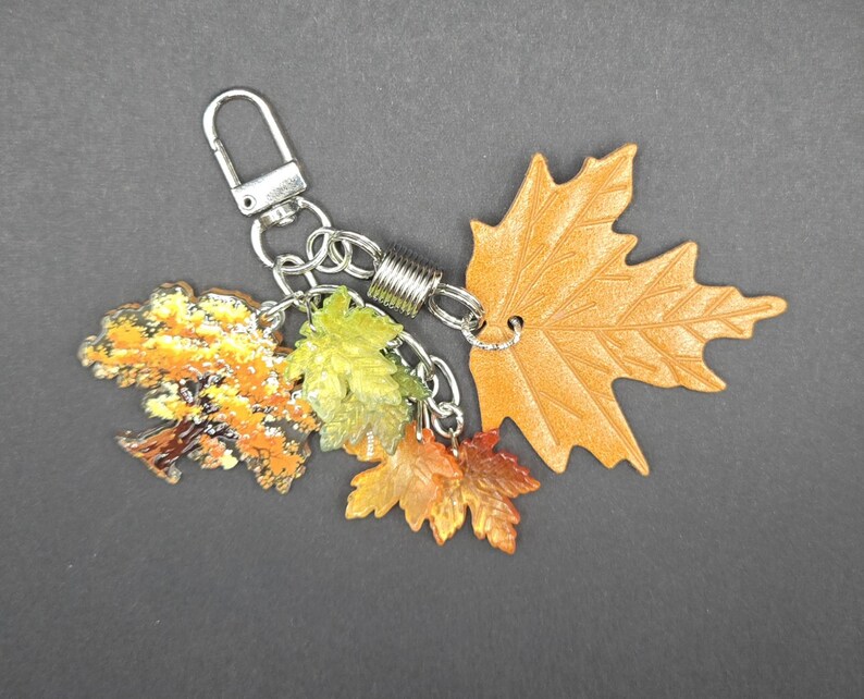 May include: A keychain featuring a silver-toned metal lobster clasp and a cylindrical connector attached to four distinct leaf-shaped charms. The charms include a large tan textured leaf, a small green leaf, a small orange leaf, and a tree-shaped charm.