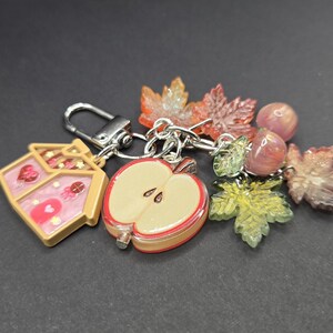 May include: A silver-toned metal keychain featuring a collection of charms including a house-shaped pendant with pink and gold accents, a halved apple charm, several translucent maple leaf charms in autumn colors, and two small round pink beads.