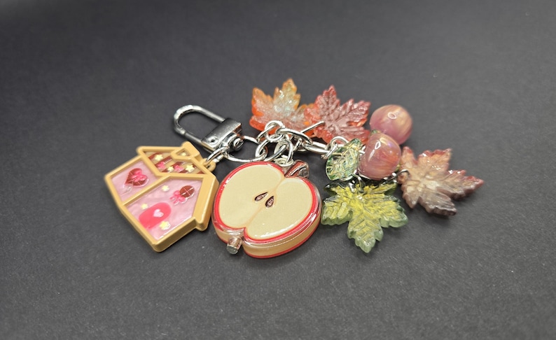 May include: A silver-toned metal keychain featuring a collection of charms including a house-shaped pendant with pink and gold accents, a halved apple charm, several translucent maple leaf charms in autumn colors, and two small round pink beads.