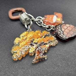 May include: A keychain featuring three charms attached to a silver-toned metal ring and a brown plastic clasp. The charms include a tree with yellow and orange autumn leaves, a brown acorn with a textured cap, and a small orange and white fox.
