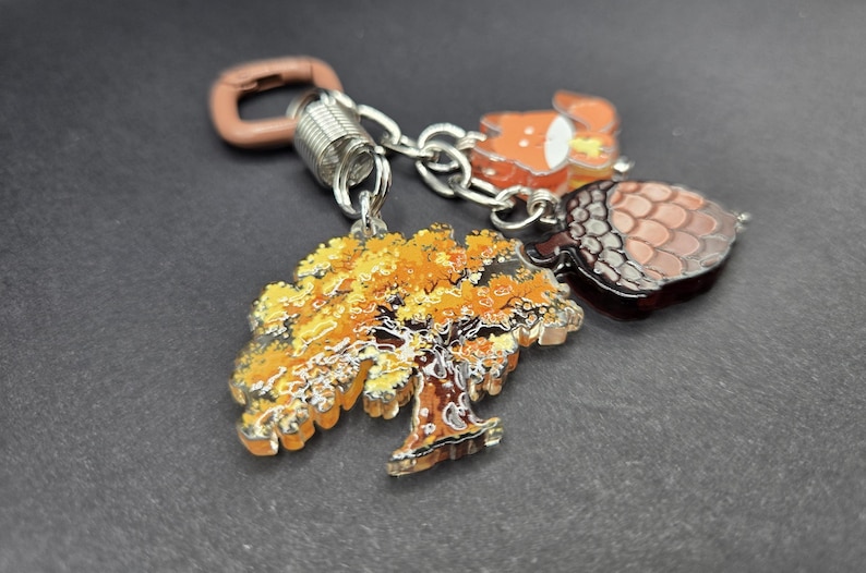 May include: A keychain featuring three charms attached to a silver-toned metal ring and a brown plastic clasp. The charms include a tree with yellow and orange autumn leaves, a brown acorn with a textured cap, and a small orange and white fox.