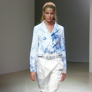 Peut inclure: Une femme porte une jupe blanche et une veste bleu clair avec un motif bleu et blanc. La veste est boutonnée et a des manches longues. Une ceinture bleu clair cintre la taille. La femme porte des sandales blanches.