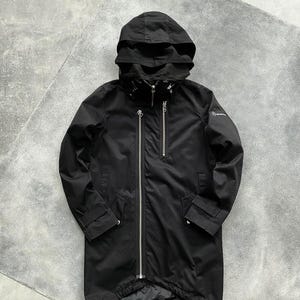 May include: A black parka with a hood and a full-length zip. The jacket has a drawstring hood, long sleeves with buttoned cuffs, and a longer back hem. The parka is made of a smooth, water-resistant material, suitable for wet weather.