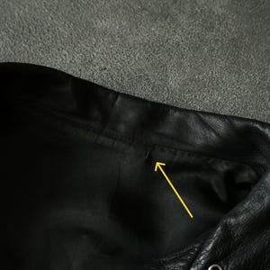 May include: Close-up of a black leather garment, possibly a jacket or vest, showing the collar and a portion of the front. The leather has a textured appearance, with visible stitching and metal eyelets.