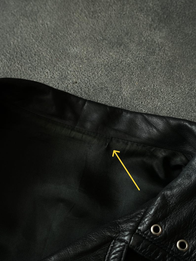 May include: Close-up of a black leather garment, possibly a jacket or vest, showing the collar and a portion of the front. The leather has a textured appearance, with visible stitching and metal eyelets.