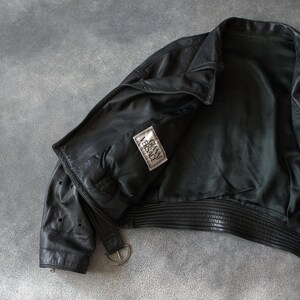 May include: Black leather jacket with a ribbed hem and a silver-tone buckle. The jacket features a "GIANNI VERSACE" label. The jacket is open, showing the interior lining.