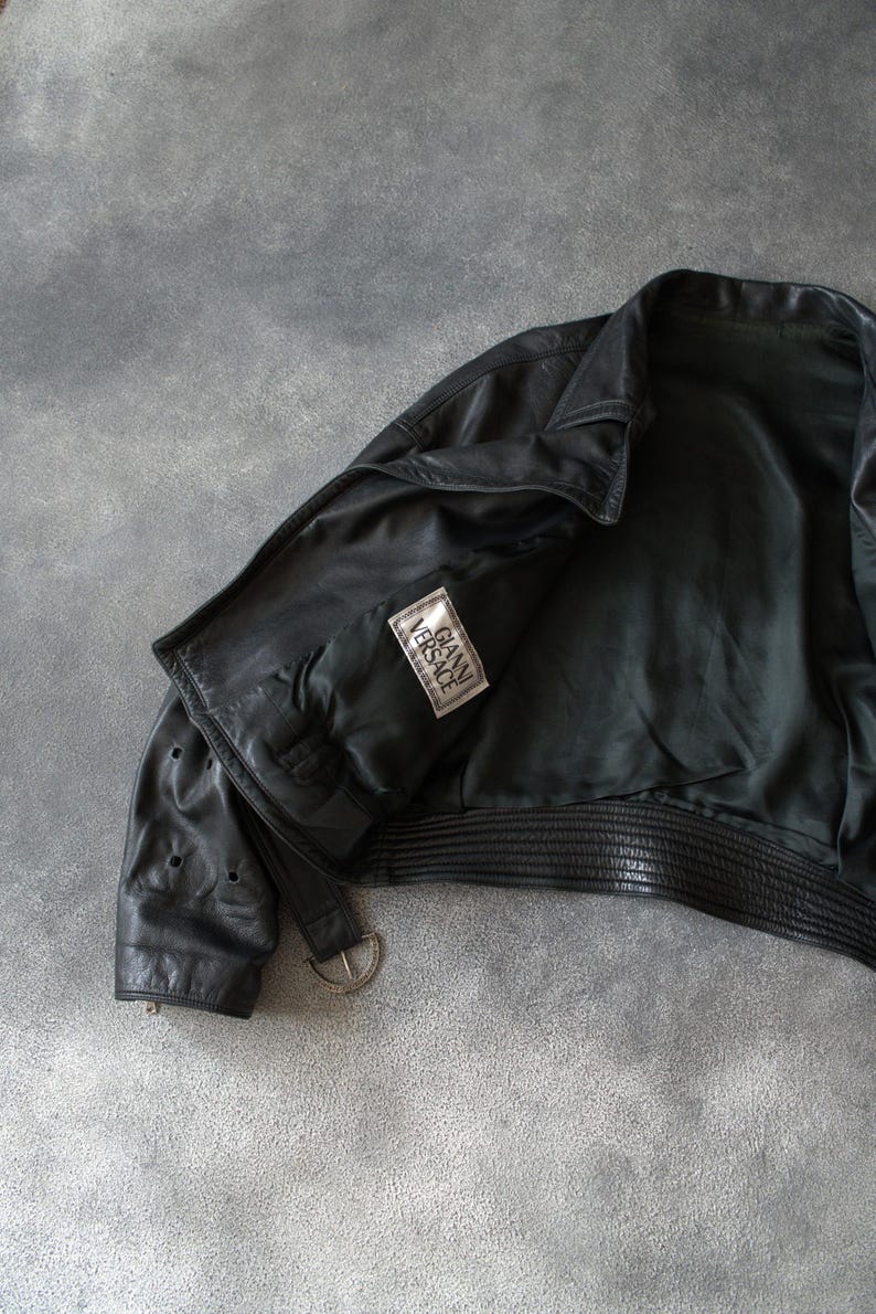 May include: Black leather jacket with a ribbed hem and a silver-tone buckle. The jacket features a "GIANNI VERSACE" label. The jacket is open, showing the interior lining.