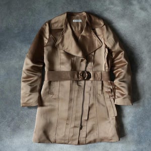 May include: A tan-colored trench coat with a matching belt and buckle. The coat has a classic design with a notched collar, long sleeves, and a button closure. The fabric has a satin sheen, and the coat is displayed on a gray background.