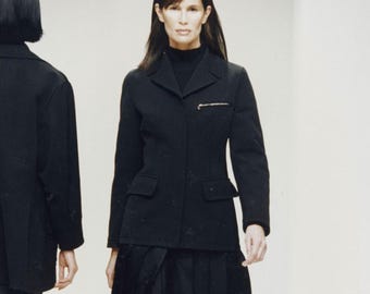 Könnte beinhalten: Ein schwarzer Blazer mit Reverskragen, zwei Vordertaschen und einem Reißverschlussdetail auf der Brust. Der Blazer wird mit einem schwarzen Faltenrock kombiniert. Eine Person trägt das Outfit.