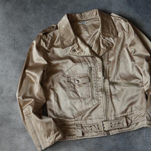 Op de afbeelding: Een beige jasje in bikerstijl met een glanzende afwerking. De jas heeft een kraag, meerdere ritszakken en een riem in de taille. De jas ligt plat op een grijze ondergrond. De merknaam is zichtbaar.