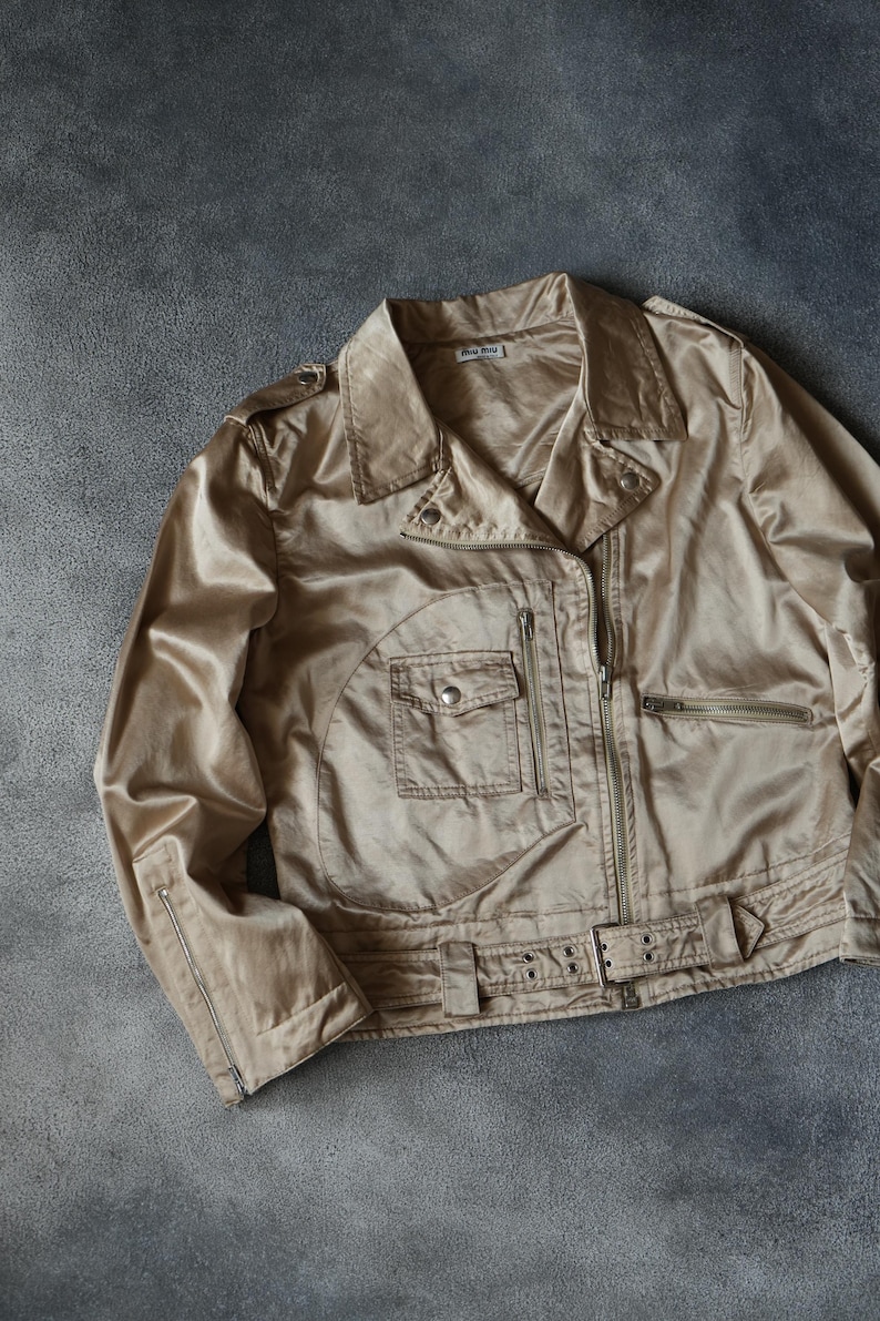 Op de afbeelding: Een beige jasje in bikerstijl met een glanzende afwerking. De jas heeft een kraag, meerdere ritszakken en een riem in de taille. De jas ligt plat op een grijze ondergrond. De merknaam is zichtbaar.