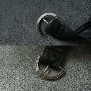 May include: A close-up shot of a black leather belt with a silver buckle. The buckle is a half-circle design with a single prong. The leather belt is thick and appears to be of good quality. The background is a textured grey surface.