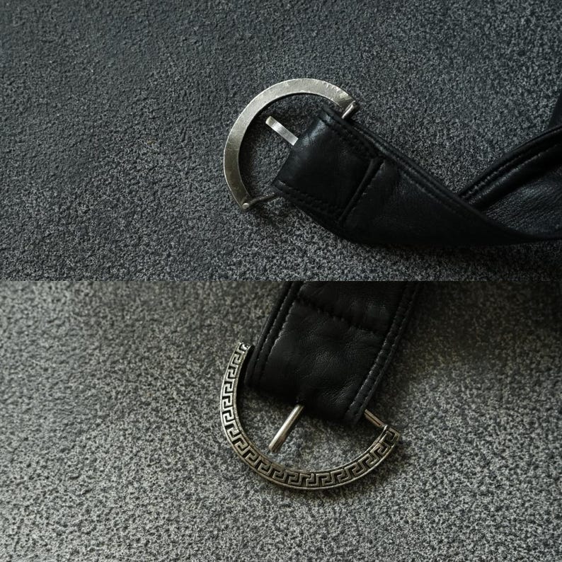 May include: A close-up shot of a black leather belt with a silver buckle. The buckle is a half-circle design with a single prong. The leather belt is thick and appears to be of good quality. The background is a textured grey surface.