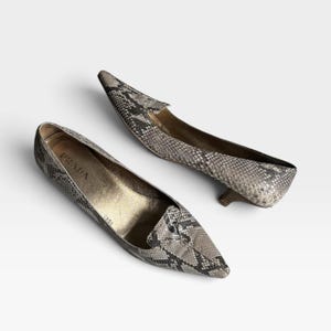 May include: A pair of pointed-toe kitten heel pumps featuring a grey and black snakeskin pattern. The interior of the shoes is lined in a metallic gold tone, with the brand name Prada printed in black on the inner sole of the left shoe.