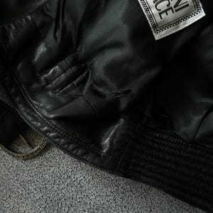 May include: Close-up of a black leather jacket with a Gianni Versace label. The jacket features a quilted design on the lower hem and a silver-toned buckle. The interior lining is visible, showcasing the quality craftsmanship.