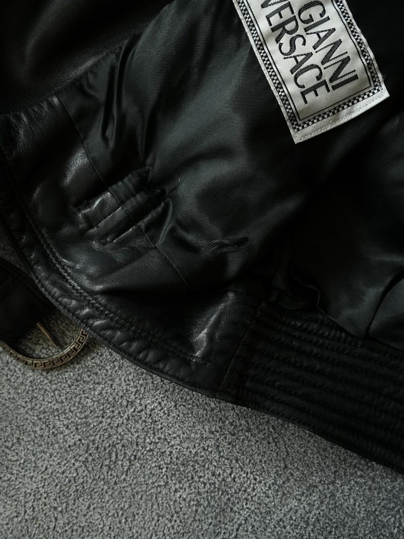 May include: Close-up of a black leather jacket with a Gianni Versace label. The jacket features a quilted design on the lower hem and a silver-toned buckle. The interior lining is visible, showcasing the quality craftsmanship.