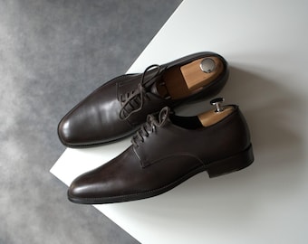Könnte beinhalten: Ein Paar dunkelbraune Lederschuhe mit Schnürsenkeln, präsentiert auf einer weißen Oberfläche. Schuhspanner aus Holz befinden sich in den Schuhen. Die Schuhe haben ein klassisches, poliertes Aussehen.