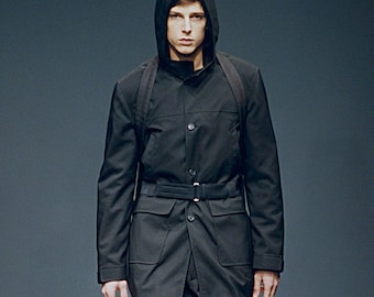Könnte beinhalten: Schwarze Kapuzenjacke mit Gürtel und großen Taschen. Die Jacke hat einen Knopfverschluss und eine hochgezogene Kapuze. Das Model trägt die Jacke und schaut in die Kamera.