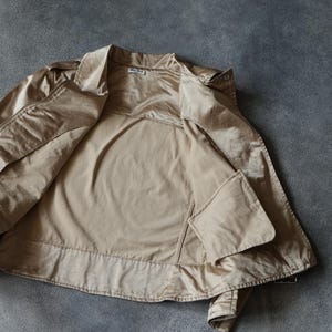 Op de afbeelding: Een beige jasje met een glanzende, satijnachtige afwerking. De jas is open, waardoor de binnenvoering en zakken zichtbaar zijn. De kraag is omgeslagen en een klein label is zichtbaar bij de nek. De jas ligt plat op een grijze ondergrond.