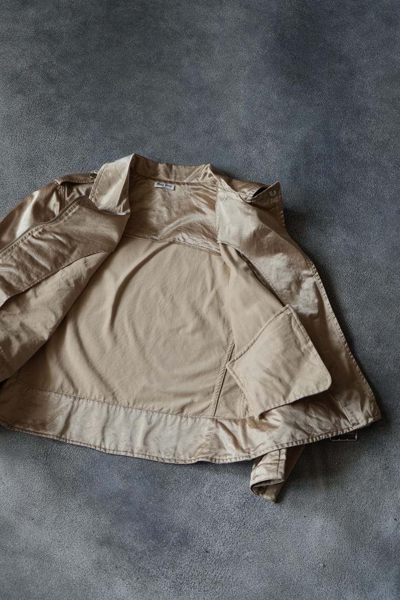 Op de afbeelding: Een beige jasje met een glanzende, satijnachtige afwerking. De jas is open, waardoor de binnenvoering en zakken zichtbaar zijn. De kraag is omgeslagen en een klein label is zichtbaar bij de nek. De jas ligt plat op een grijze ondergrond.