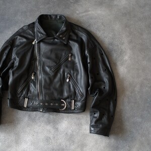 May include: Black leather jacket featuring an asymmetrical zip, multiple zipped pockets, and a belted waist. The jacket has a classic biker style with a notched collar and silver hardware.