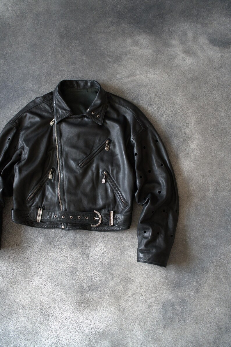 May include: Black leather jacket featuring an asymmetrical zip, multiple zipped pockets, and a belted waist. The jacket has a classic biker style with a notched collar and silver hardware.