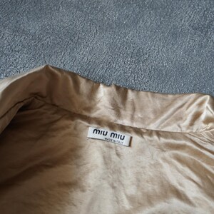 Op de afbeelding: Close-up van een beige satijnen kledingstuk met een kraag. Een wit label met de merknaam "miu miu" en "MADE IN ITALY" is zichtbaar. De stof heeft een subtiele glans.