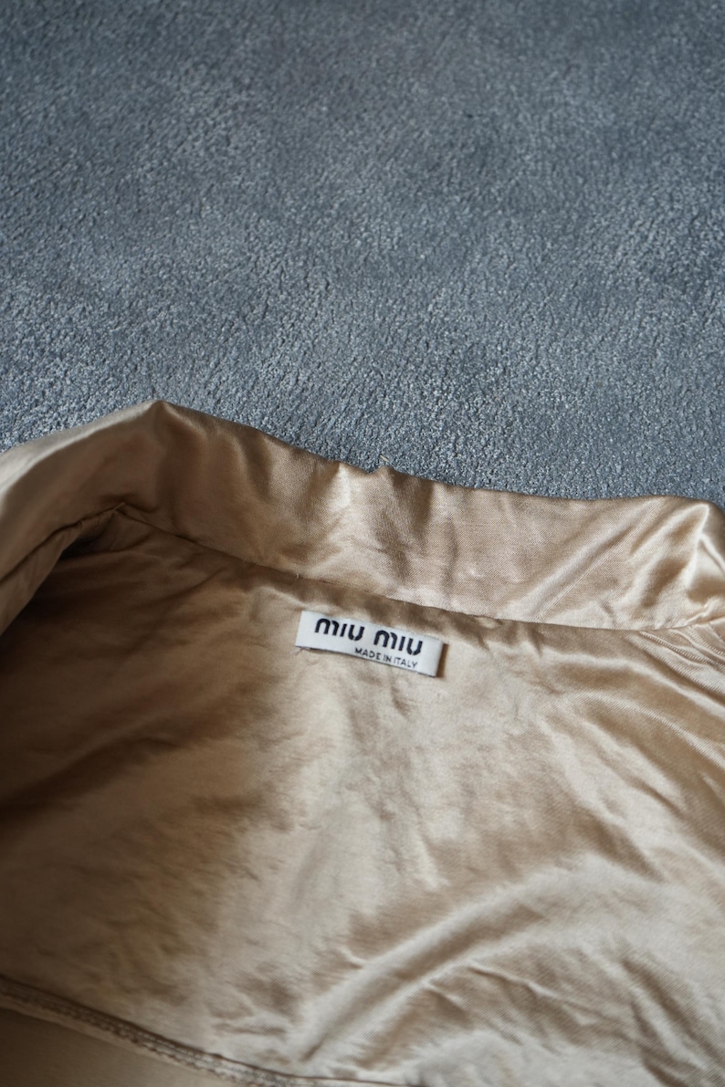Op de afbeelding: Close-up van een beige satijnen kledingstuk met een kraag. Een wit label met de merknaam "miu miu" en "MADE IN ITALY" is zichtbaar. De stof heeft een subtiele glans.