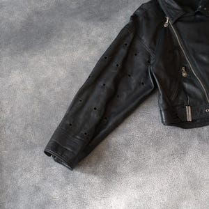 May include: Black leather jacket with a diagonal zip and a notched collar. The sleeve has multiple small, evenly spaced circular cutouts. A classic jacket style, suitable for various occasions. The jacket is made of leather.