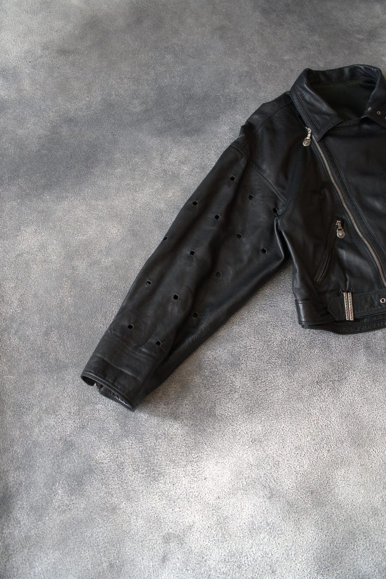 May include: Black leather jacket with a diagonal zip and a notched collar. The sleeve has multiple small, evenly spaced circular cutouts. A classic jacket style, suitable for various occasions. The jacket is made of leather.