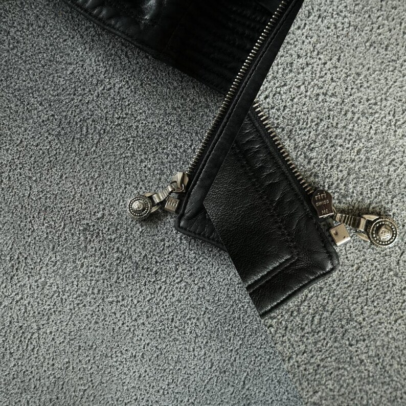 May include: Close-up of a black leather jacket with a silver zip. The zip is partially open, revealing the texture of the leather and the intricate design of the zip pull. The jacket appears to be new.