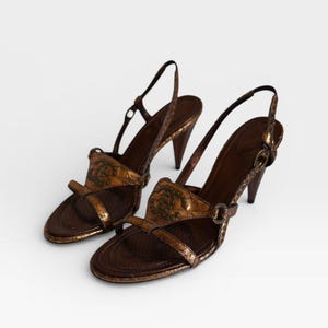 Puede incluir: Un par de sandalias de tacón de cuero marrón. Las sandalias presentan un diseño decorativo en las correas y una correa trasera. Los tacones miden aproximadamente 7,6 cm de alto.