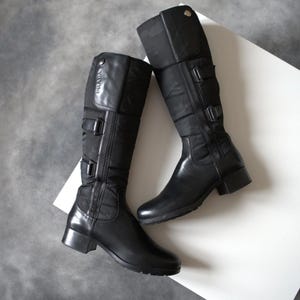 Pode incluir: Um par de botas pretas até ao joelho, combinando couro e nylon. Apresentam fivelas, fecho de correr e a marca "PRADA". As botas, com salto baixo, são exibidas numa superfície branca, realçando o design.