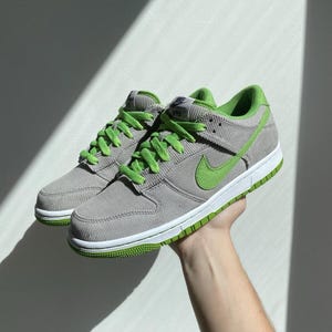 May include: A pair of gray corduroy sneakers with bright green accents. The shoes feature green laces, a green Nike swoosh, and a green sole. The sneakers are held up by a person's hand.