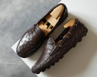 Könnte beinhalten: Ein Paar dunkelbraune Leder-Loafer mit strukturierter Oberfläche, jeweils mit einem hölzernen Schuhspanner. Die Schuhe haben ein klassisches Design mit sichtbaren Nähten und dekorativen Ösen. Auf einer weißen Oberfläche vor grauem Hintergrund.