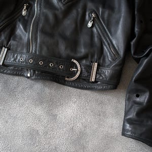 May include: A close-up shot of a black leather jacket featuring silver zippers and a decorative belt. The jacket has multiple pockets and a buckle closure. The leather is textured and the jacket is partially unzipped.