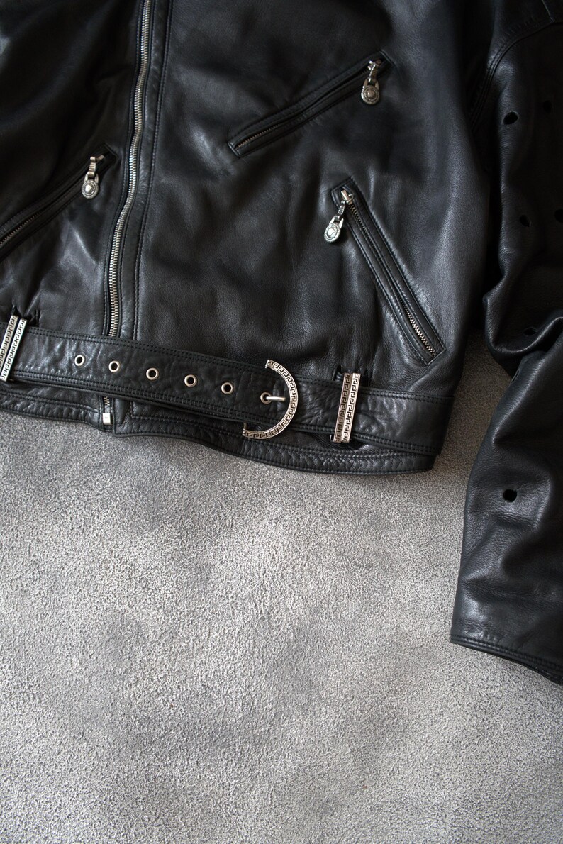 May include: A close-up shot of a black leather jacket featuring silver zippers and a decorative belt. The jacket has multiple pockets and a buckle closure. The leather is textured and the jacket is partially unzipped.