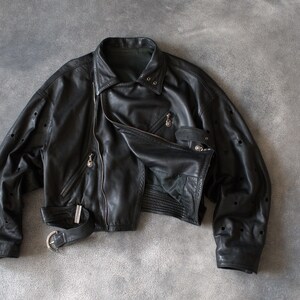 May include: Black leather jacket with an asymmetrical zip, a notched collar, and a belted waist. The jacket has multiple zipped pockets and decorative perforations on the sleeves. The jacket is a cropped style.