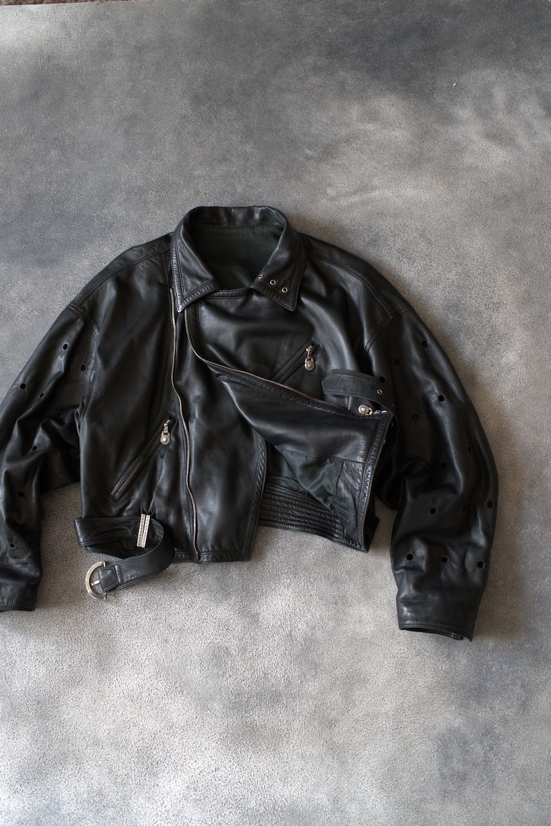 May include: Black leather jacket with an asymmetrical zip, a notched collar, and a belted waist. The jacket has multiple zipped pockets and decorative perforations on the sleeves. The jacket is a cropped style.