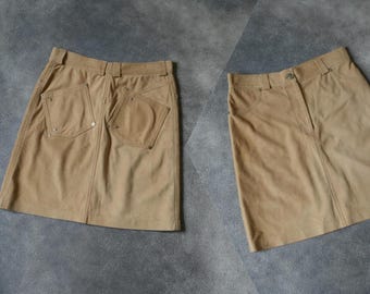 Könnte beinhalten: Zwei hellbraune Wildleder-Röcke. Ein Rock ist von hinten zu sehen, mit zwei großen Taschen und dekorativen Nähten. Der andere Rock ist von vorne zu sehen, mit Knopfverschluss und Taschen. Die Röcke sind neutralfarben.